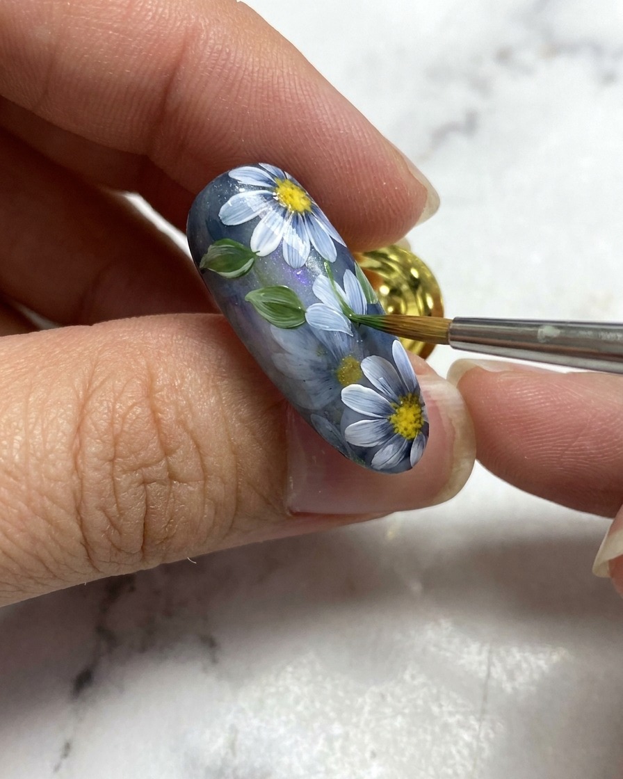 Un pennello da nail art aggiunge una piccola foglia verde accanto ai dettagli floreali bianchi su una base per unghie grigia.