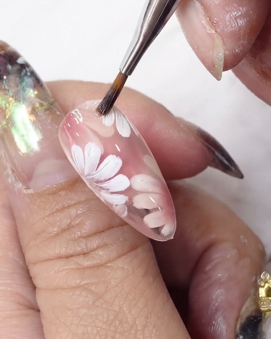Un pennello per gel che fa scorrere una mano di smalto rosa trasparente sopra i fiori bianchi dipinti su ununghia a mandorla.