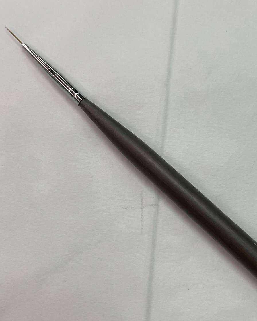 Un pennello liner fine per nail art posizionato vicino a uno schizzo di griglia guida chiaro sulla superficie di ununghia di prova.