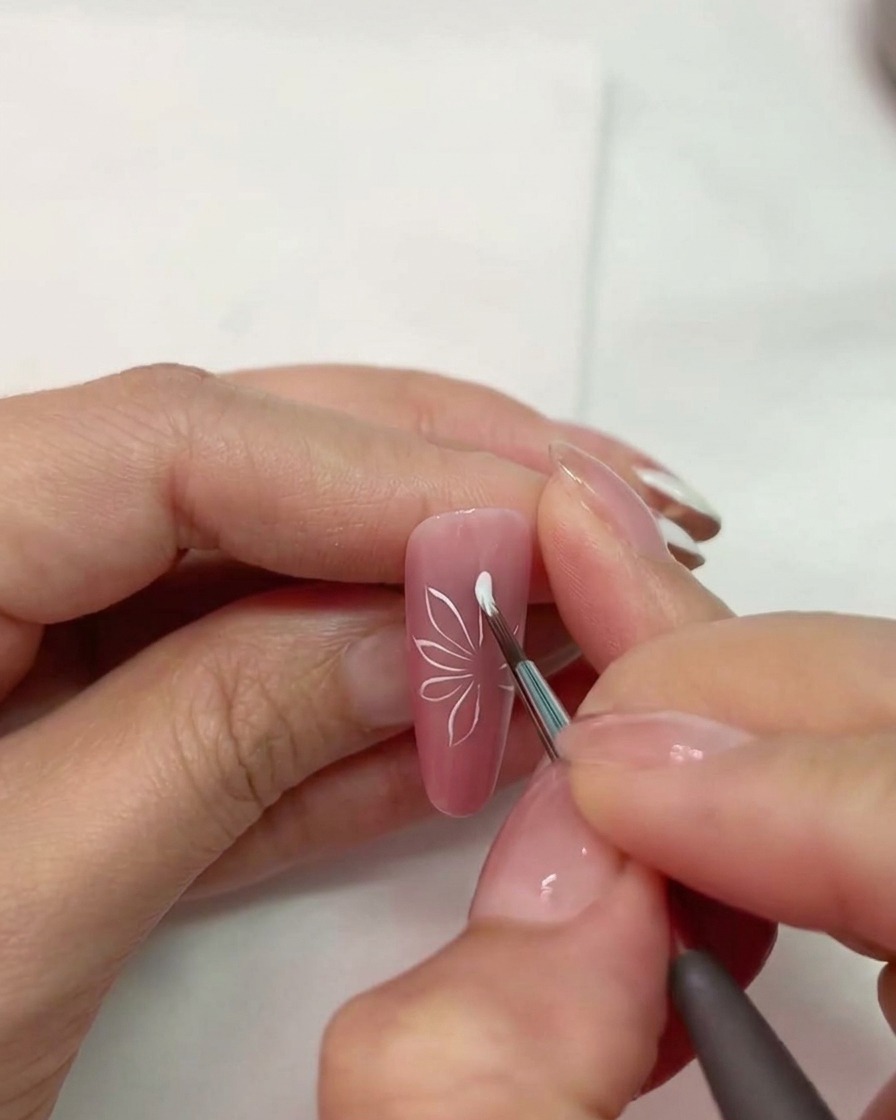 Un primo piano di un pennello per unghie che disegna forme di petali bianchi su ununghia rosa opaca.