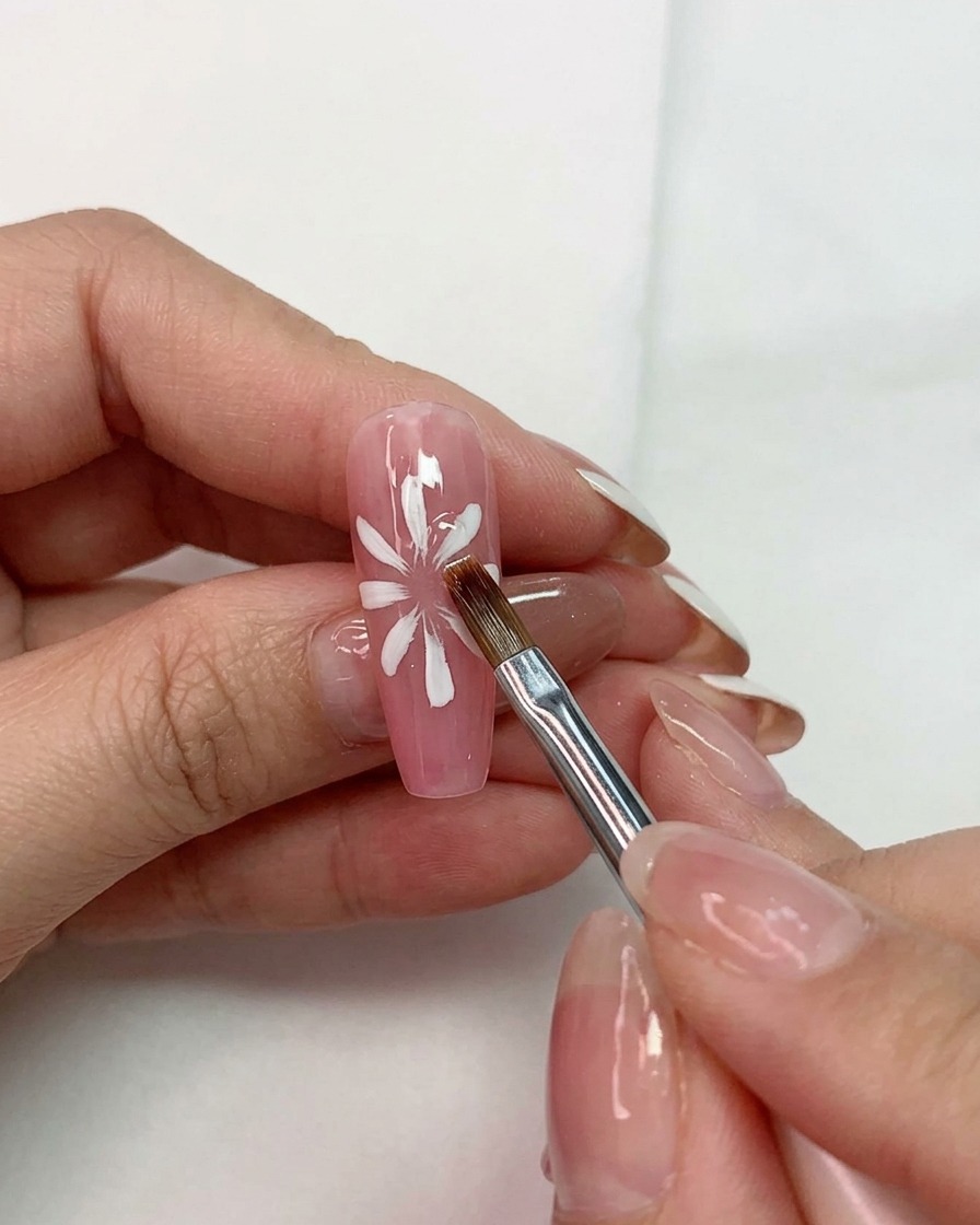 Una mano che tiene un pennello multiuso mentre modella petali bianchi su una base rosa su ununghia.