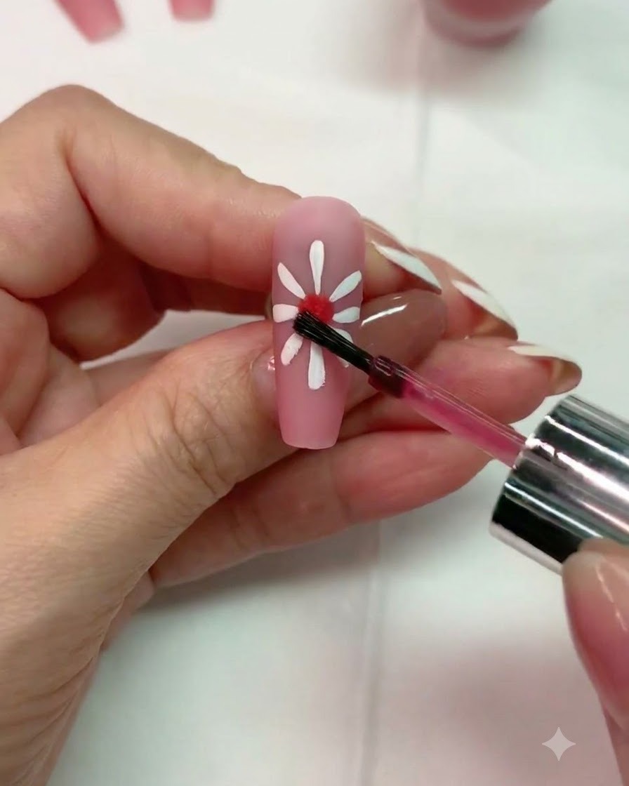Un pennello che applica una piccola goccia di blooming gel rosso al centro di un design di margherita bianca su ununghia rosa opaca.