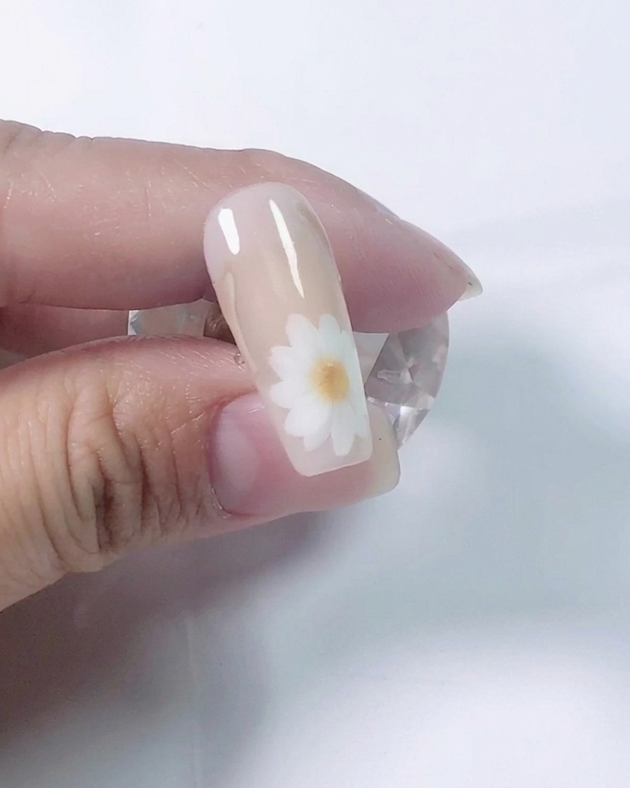 Primo piano di unonicotecnica che utilizza un pennello sottile per dipingere petali di margherita bianchi su ununghia di prova color nude.