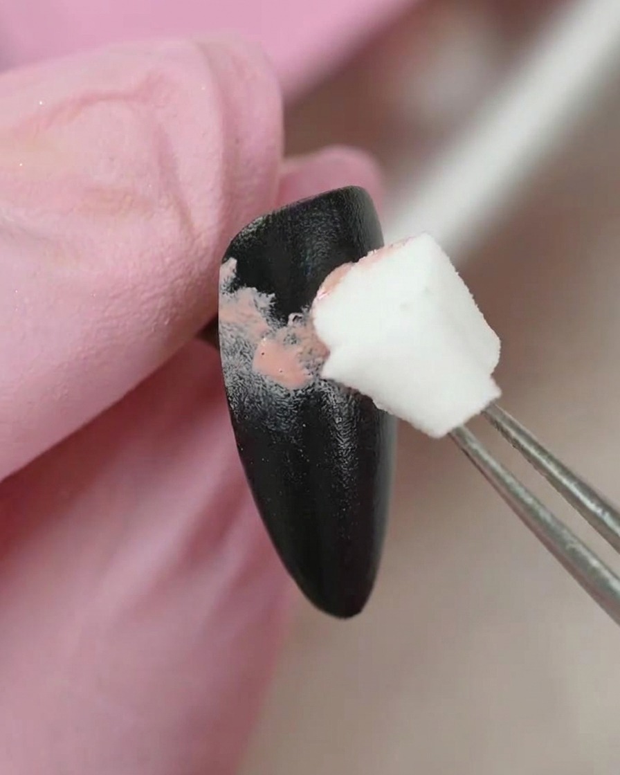 Una pinzetta che tiene un piccolo applicatore in spugna, mentre tampona delicatamente del gel colorato rosa su una superficie dunghia dipinta di nero.