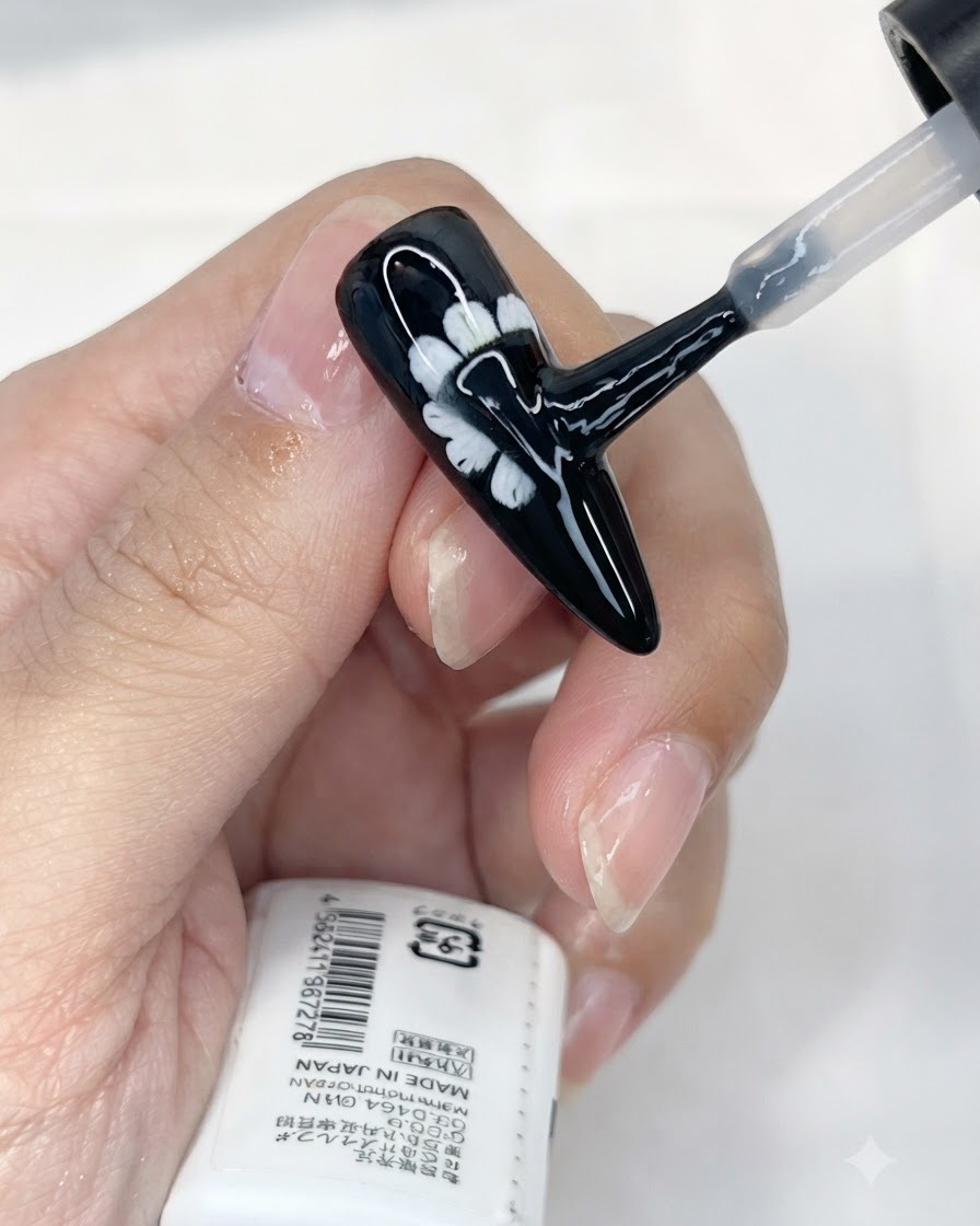 Un pennello applica uno strato di top coat opaco su ununghia nera con decorazione a margherita bianca.