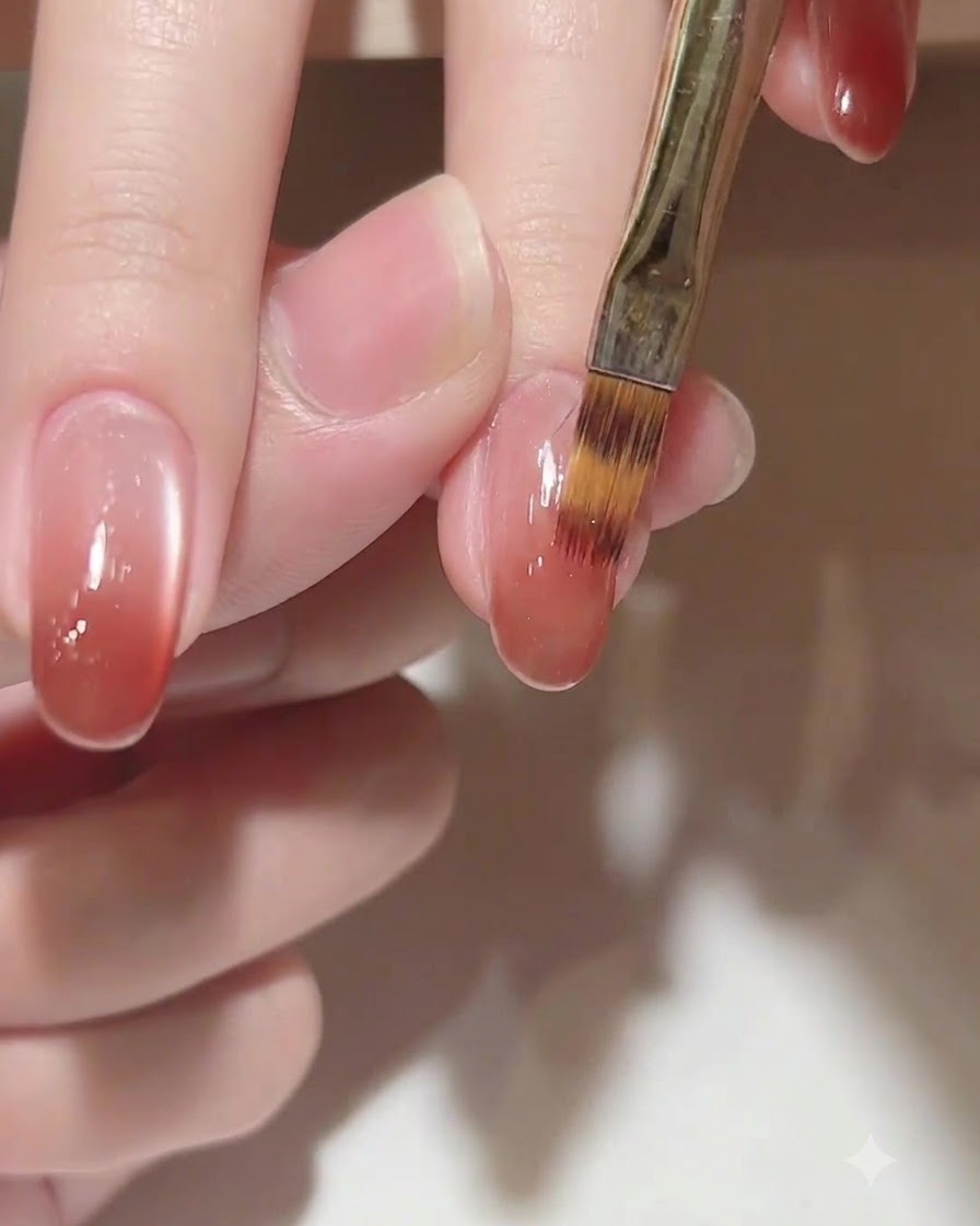 Primo piano di un pennello che sfuma delicatamente un colore gel marrone rosato caldo verso le cuticole su ununghia ovale.