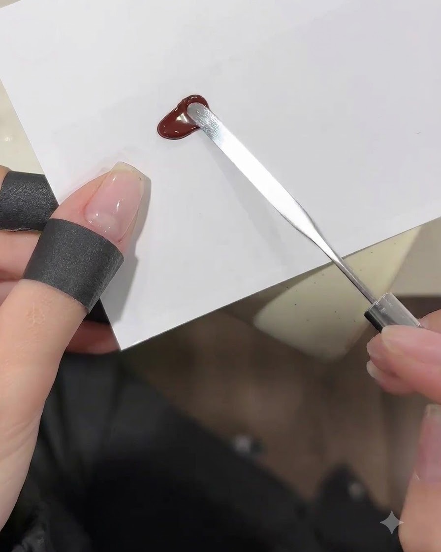 Una spatola di metallo che spalma una goccia di gel rosso vino profondo su un foglio di tavolozza bianco.