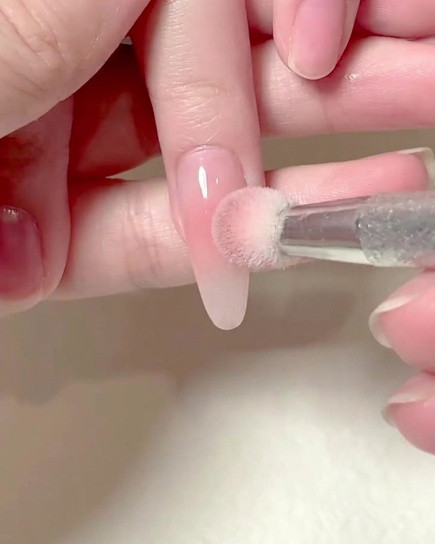 Una spugnetta quadrata tenuta da una mano che tampona delicatamente il gel rosa blush su ununghia allungata.