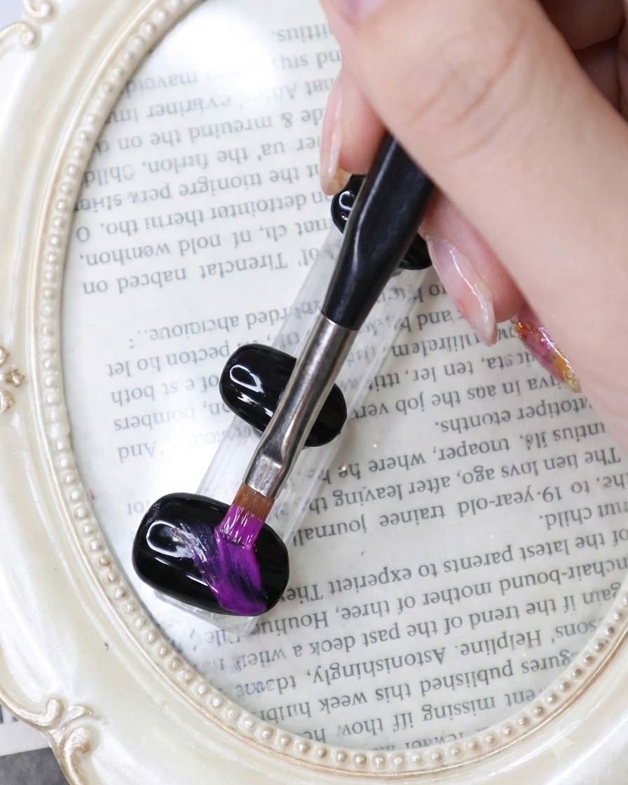 Un pennello a punta fine disegna pennellate vibranti in viola neon su ununghia in gel nero sopra uno sfondo a tema librario.