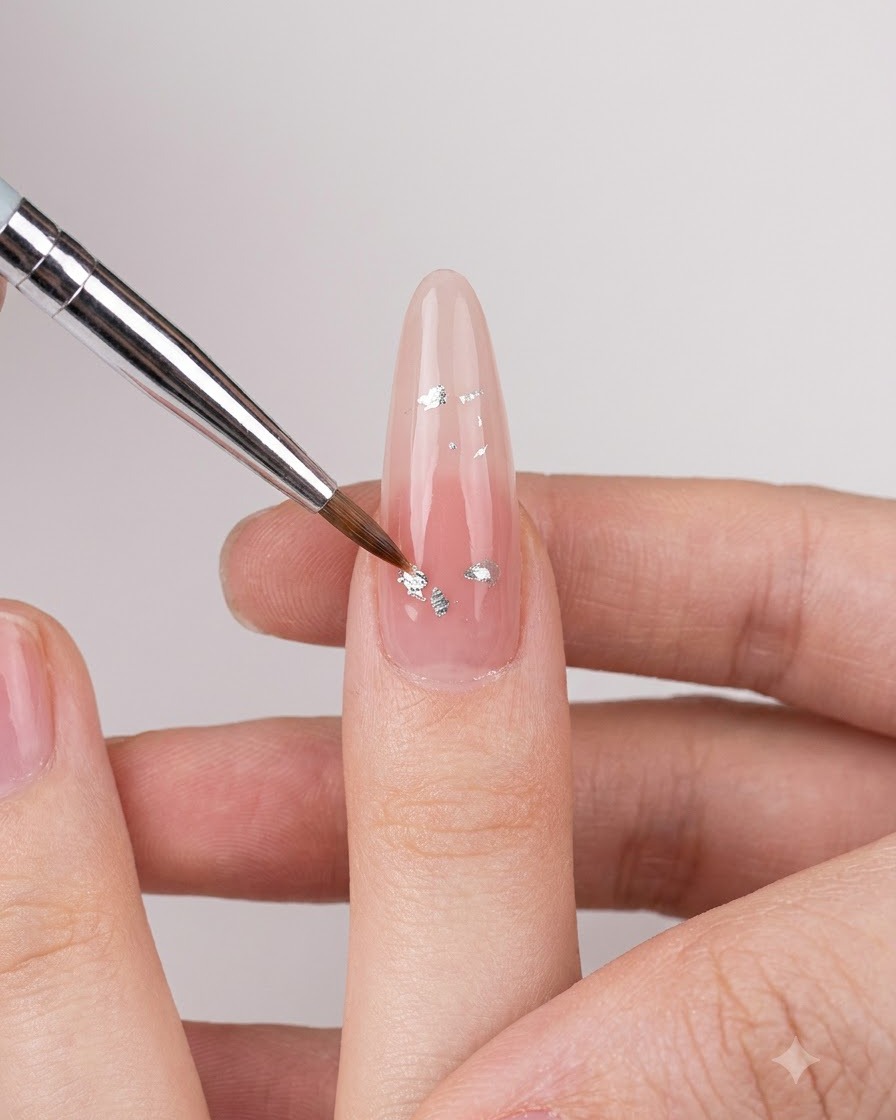 Un pennello fine per nail art posiziona piccoli fiocchi iridescenti argentati su ununghia lucida color nude.
