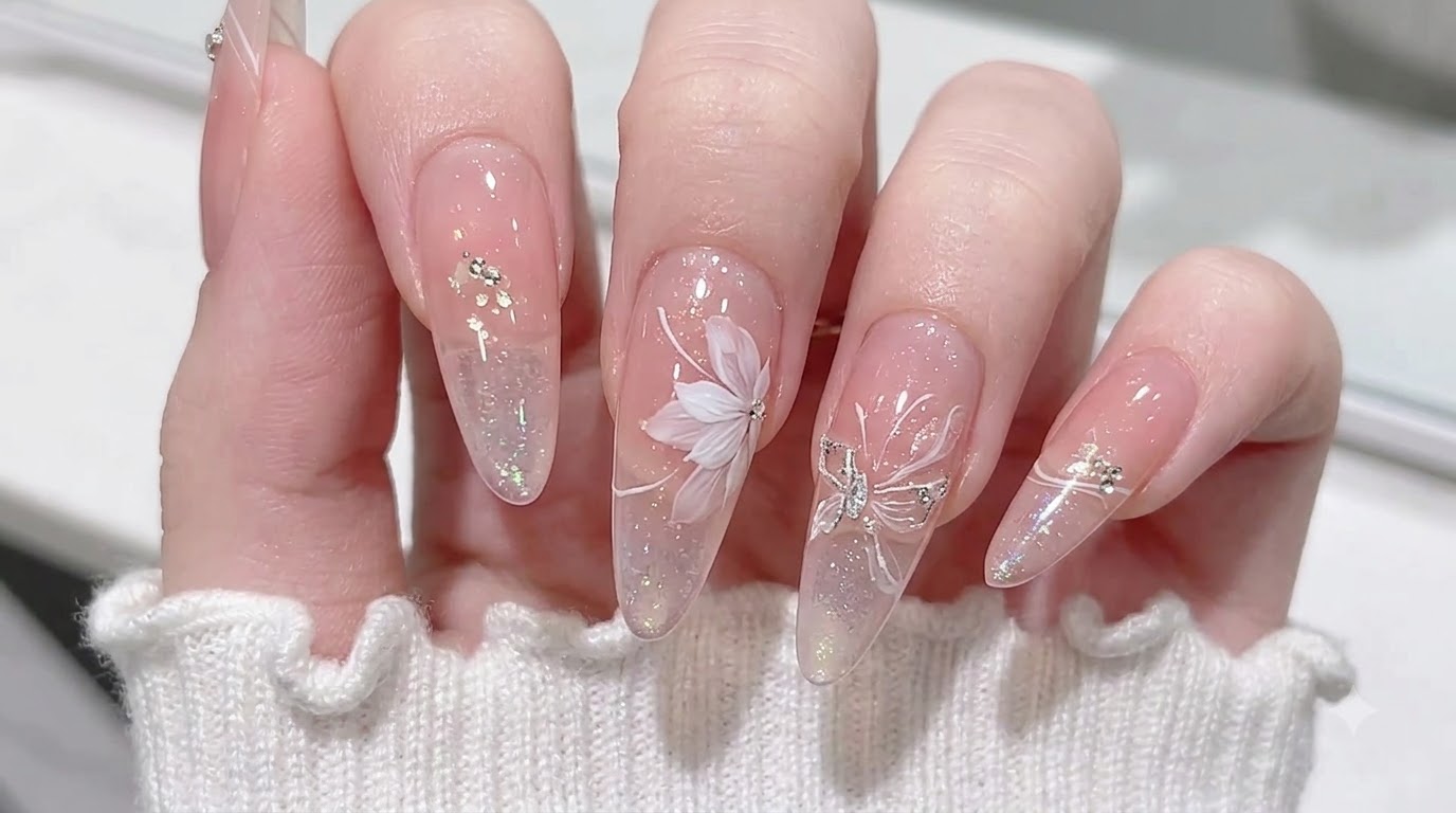 Unghie a mandorla color nude traslucido con sfumatura rosa blush, fiocchi aurora e delicata nail art floreale bianca.