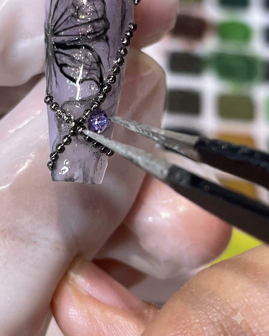Un paio di pinzette che posizionano uno strass viola a punta su una superficie ungueale decorata.