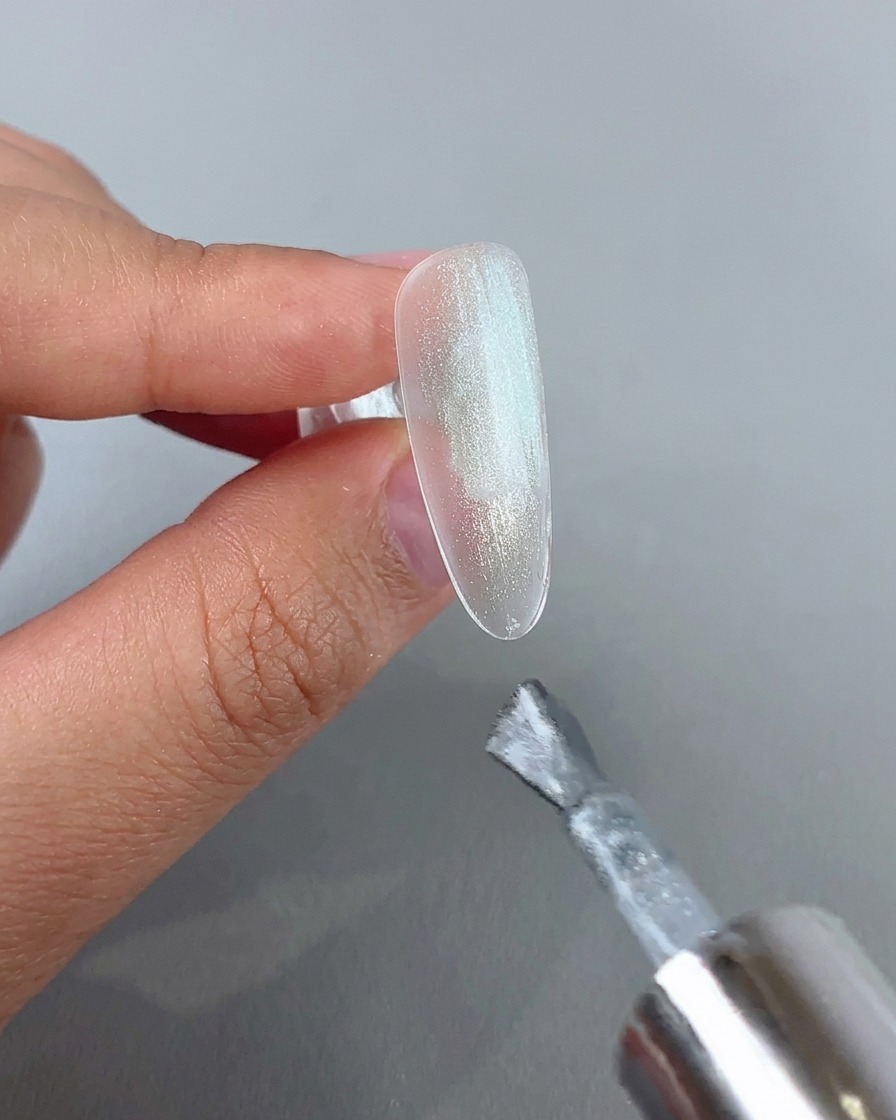 Un pennello per unghie che applica uno strato di gel cat eye argento brillante su una punta dunghia trasparente.