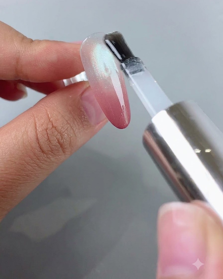 Un pennello che applica il top coat trasparente su ununghia che ha un blush sfumato rosa e un effetto cat eye argento.