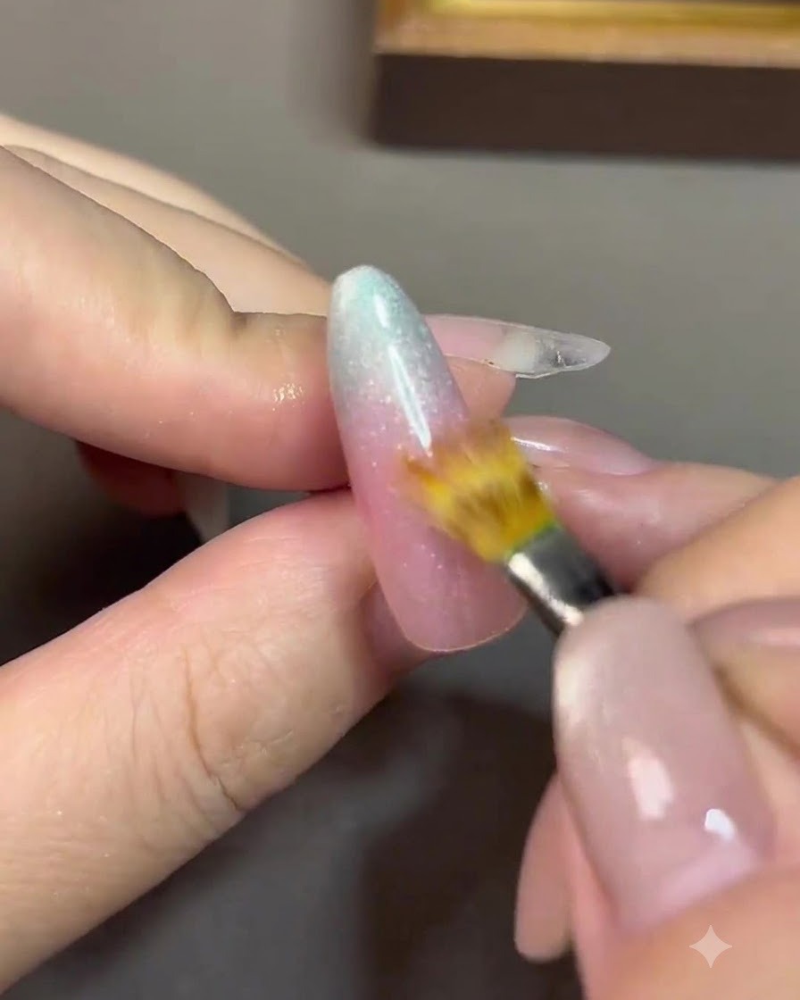 Un pennello a punta fine che sfuma la linea di transizione tra un gel nude rosato e un gel blu su ununghia.