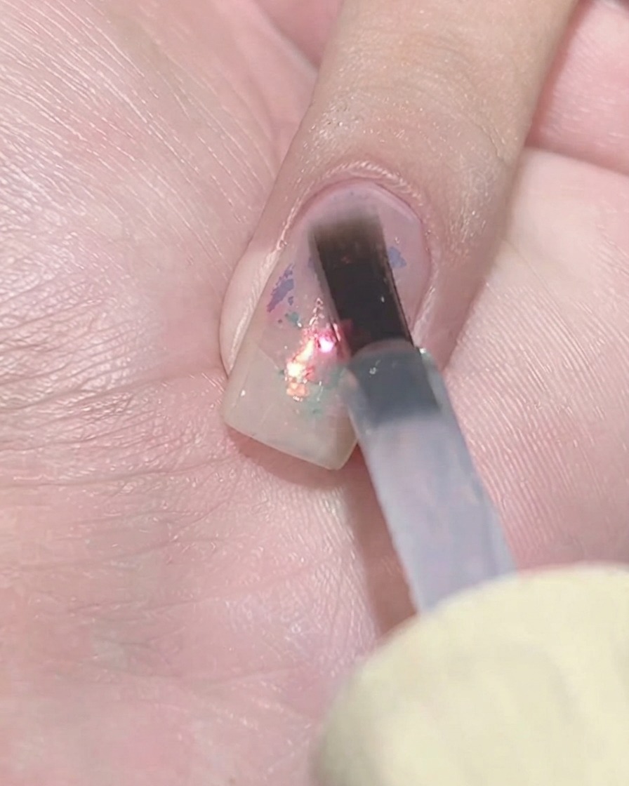 Un pennello in gel piatto che tampona uno strato di smalto viola trasparente su un cluster di fiocchi texturizzato su ununghia.