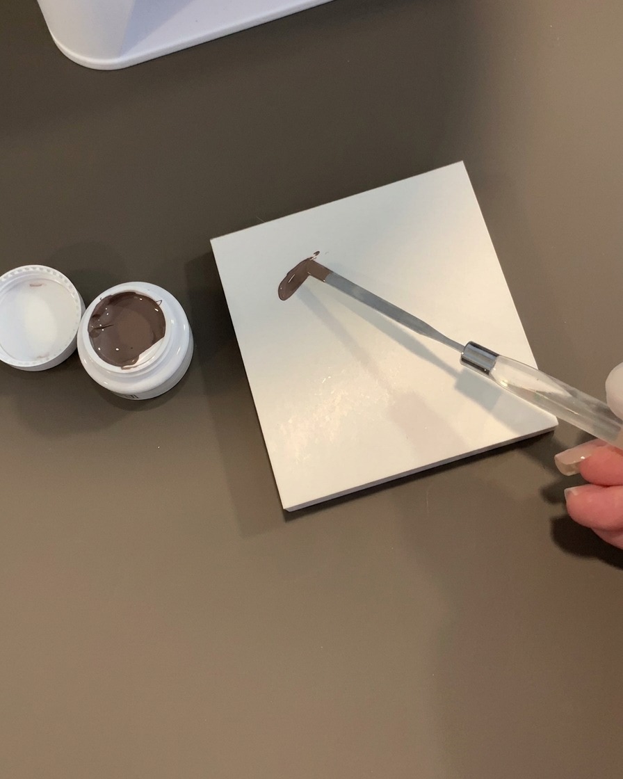 Una spatola di metallo che regge una goccia di gel grigio-marrone su una tavolozza di piastrelle quadrate bianche accanto a un barattolo aperto.