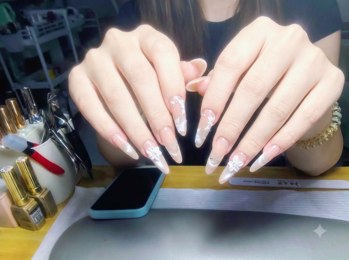 Entrambe le mani sollevate contro lo sfondo di una postazione di lavoro, che mostrano un set completo di unghie sfumate perla nude lucide con nail art floreale bianca.