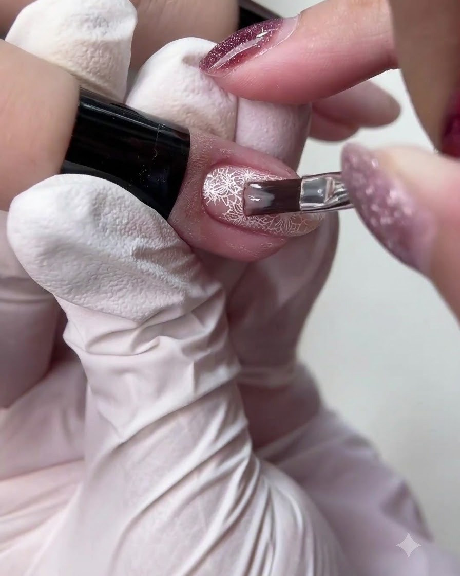 Un piccolo pennello da unghie che applica uno smalto in gel nudo traslucido sopra un dettagliato disegno di nail art in pizzo bianco.