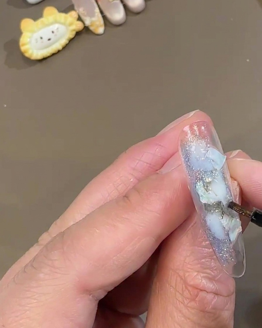 Un pennello fine per dettagli tampona polvere fatata bianca brillante su ununghia press-on trasparente decorata con pezzi di conchiglia blu.