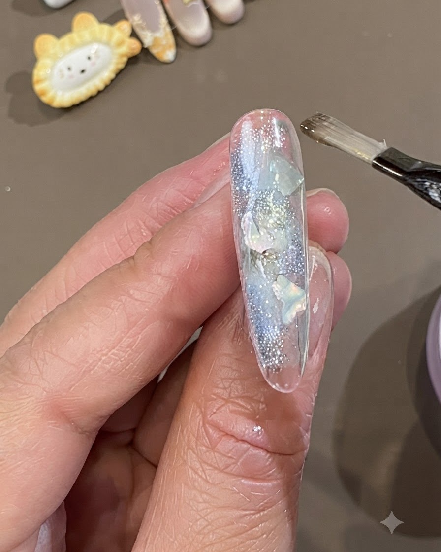 Un pennello piatto per gel applica uno strato spesso di gel di incapsulamento morbido e trasparente su ununghia decorata con pezzi di conchiglia e glitter.