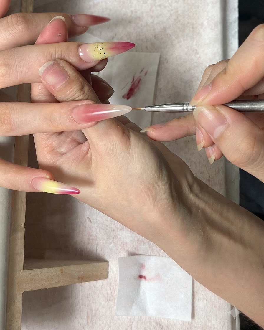 Un pennello fine per nail art che applica punti marroni come stami su ununghia sfumata.