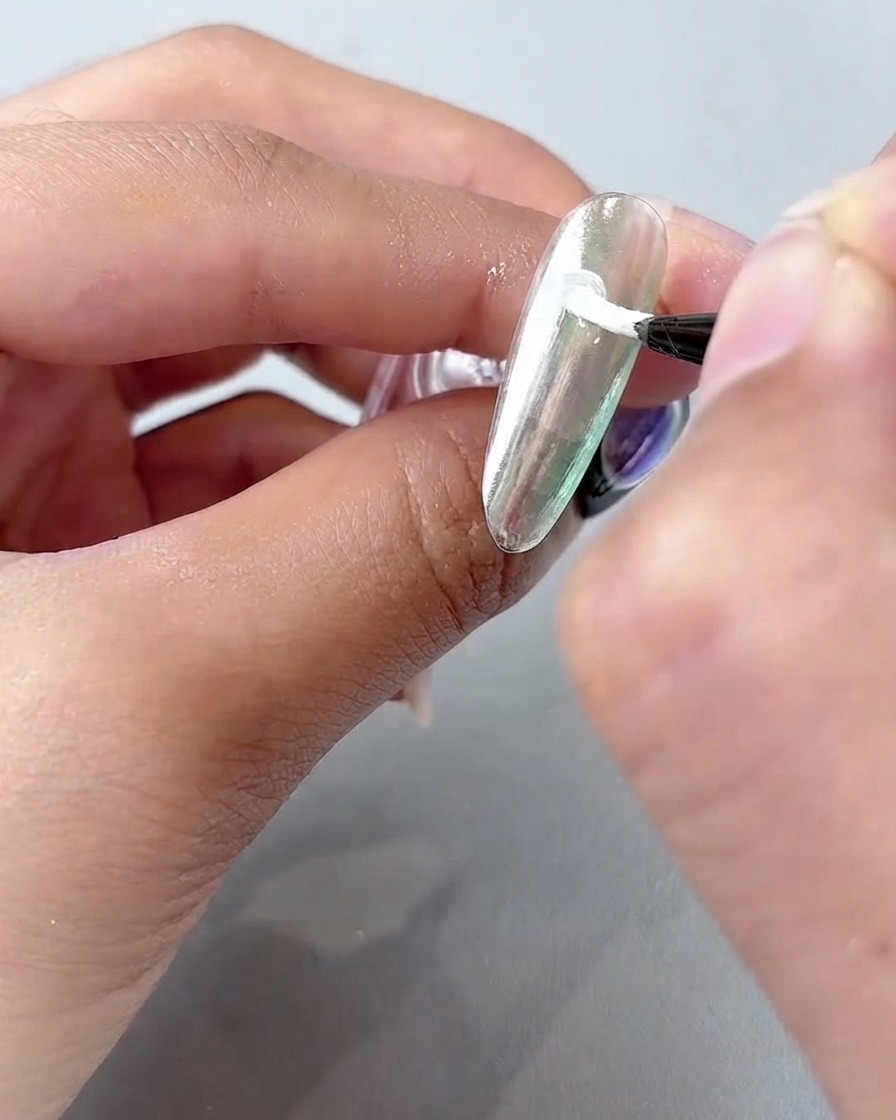 Un pennello fine per nail art che applica gel da pittura bianco in petali curvi su una superficie di unghia cromata argentata.
