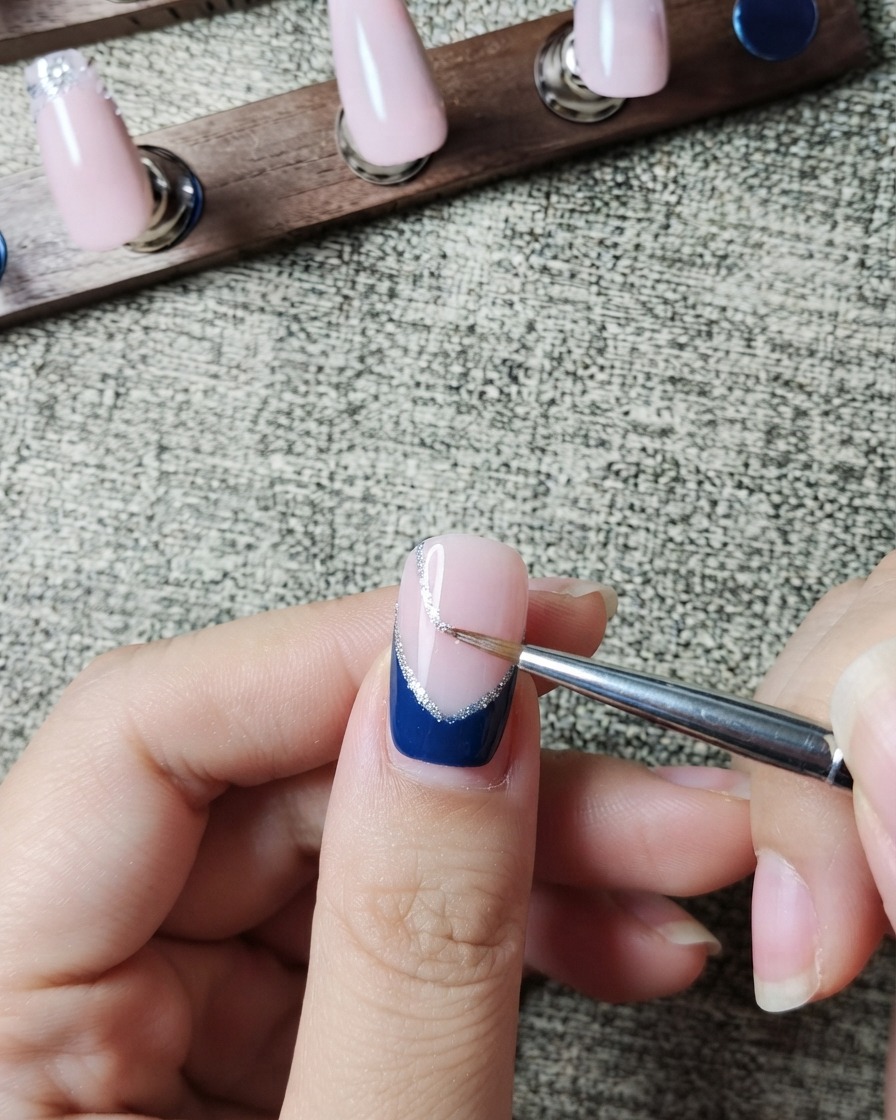 Un pennello liner che dipinge con cura una sottile linea di glitter argento lungo il margine interno di una French blu scuro.