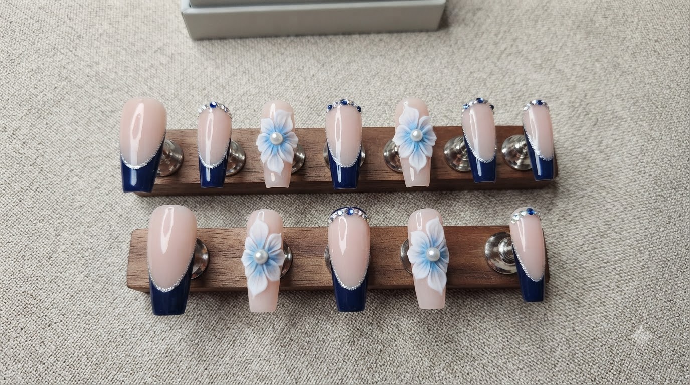 Un set completo di unghie posticce personalizzate con base nude, French blu scuro, glitter argento, strass e fiori 3D bianchi e blu.