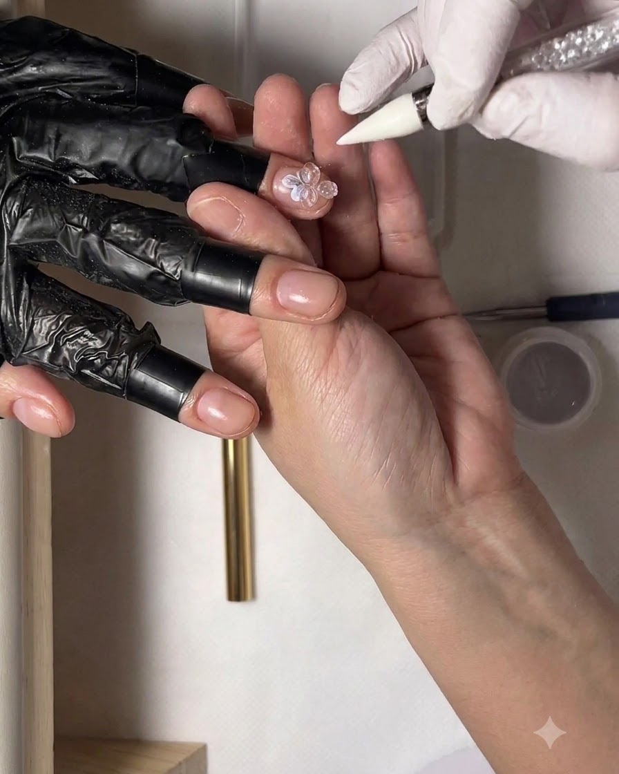 Una mano guantata che tiene una matita di cera con uno strass sulla punta, posizionandolo al centro di un design floreale per unghie.