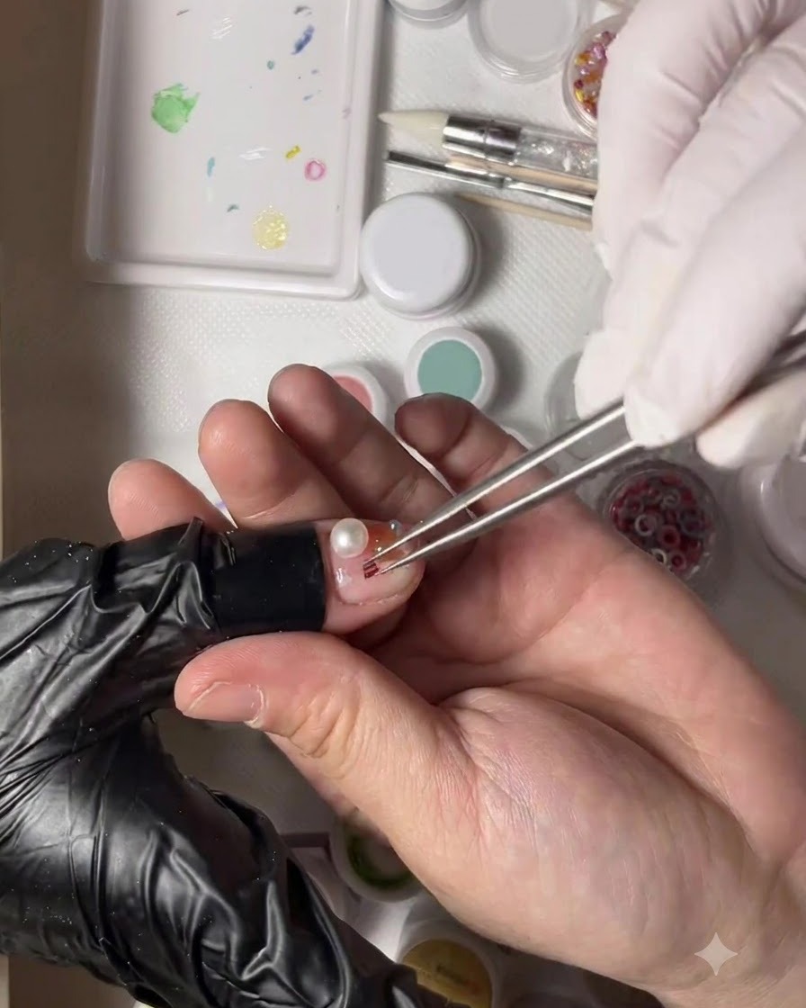 Una mano guantata che tiene le pinzette posiziona una minuscola perla doro accanto a una perla su ununghia del pollice decorata.