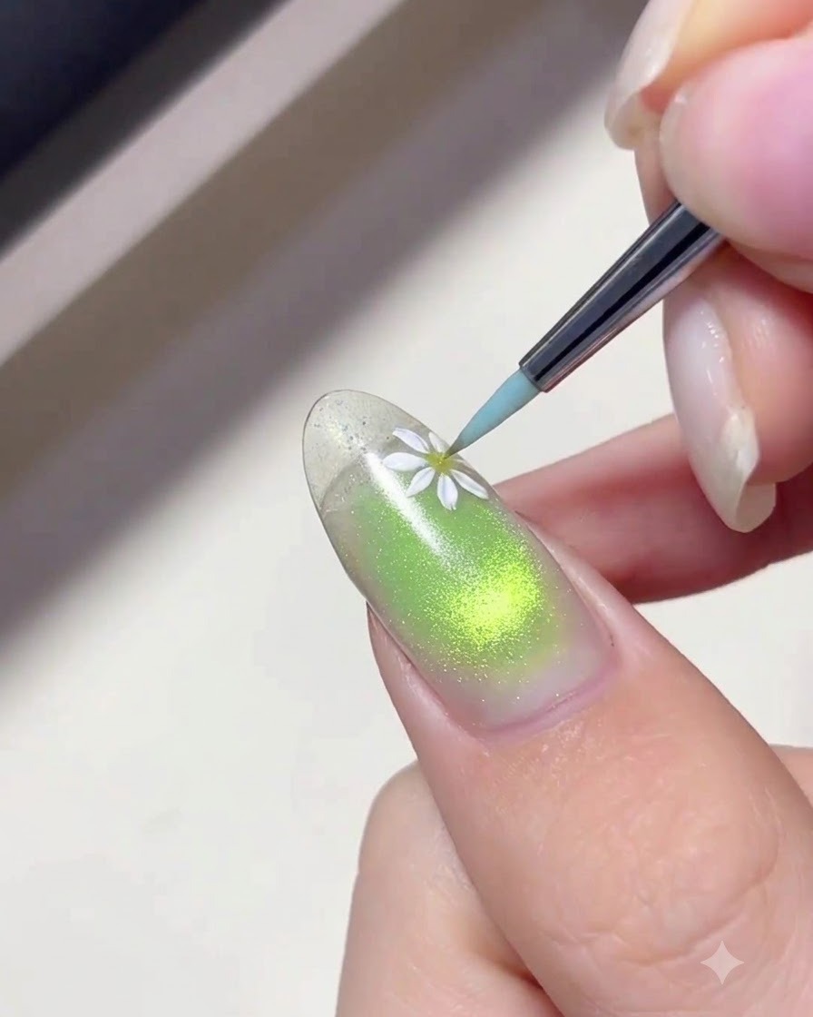 Un pennellino liner sottile applica del gel bianco per dipingere delicati petali di margherita su ununghia con sfumatura verde.