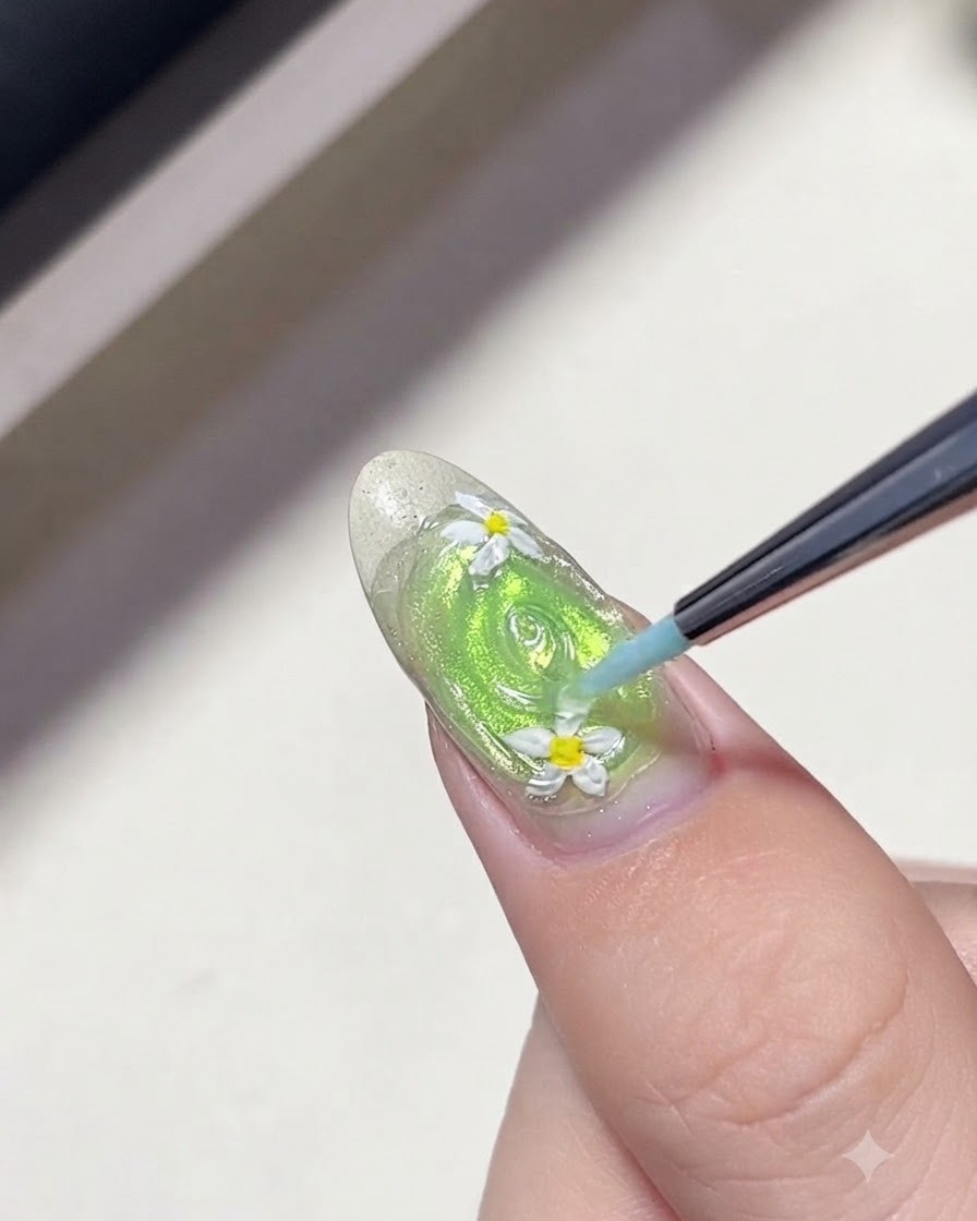 Un pennello liner sottile disegna cerchi concentrici di gel trasparente attorno ai fiori di margherita su ununghia verde per ricreare le increspature dellacqua.