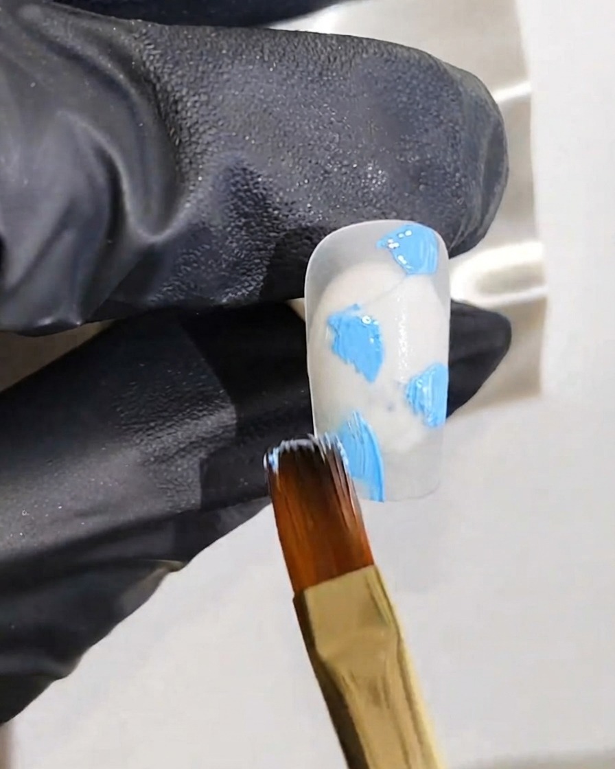 Una mano guantata che tiene un pennello fine per nail art che applica punti di smalto gel blu su una base per unghie bianca.