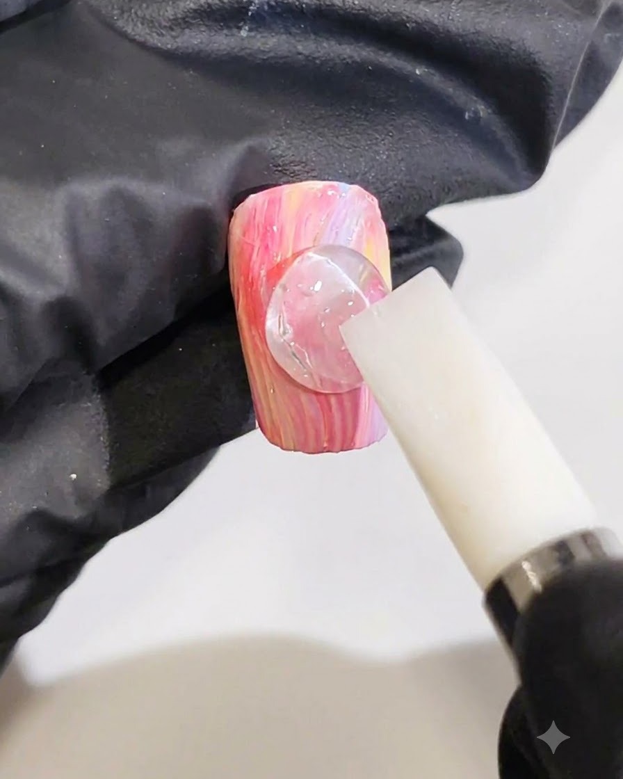 Una mano guantata che usa uno strumento in silicone bianco per modellare una goccia trasparente di gel 3D in una linea donda su ununghia marmorizzata rosa e gialla.
