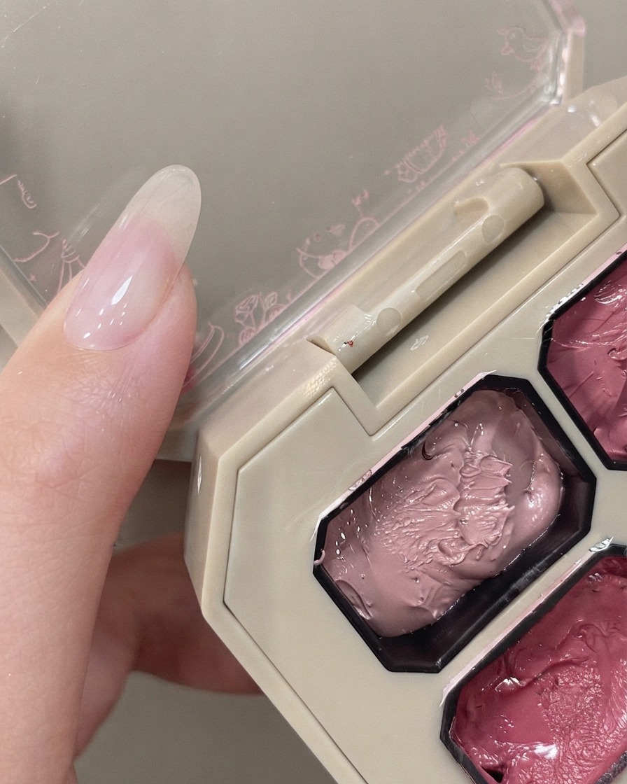 Un applicatore in spugna che fluttua sopra una palette di pigmenti rosa accanto a una mano con ununghia naturale.