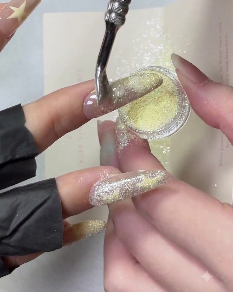 Una mano inclinata lateralmente mentre la polvere di diamante frantumato gialla viene spolverata sul bordo di una punta french manicure.